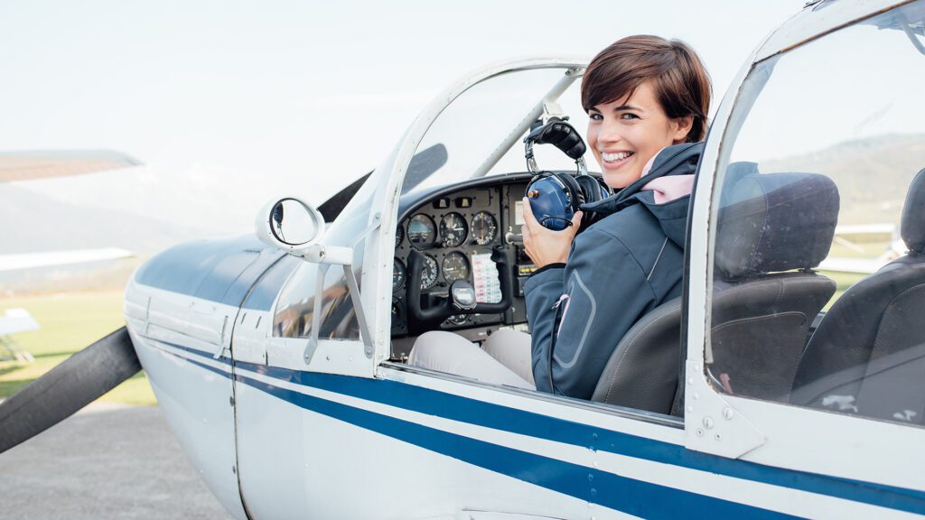 Smiling female pilot in the light aircraft cockpit, she is holding aviator headset and looking at camera
