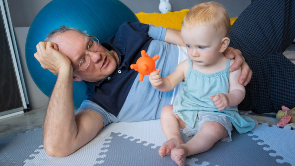 Adorable red-haired newborn sitting on floor and playing toy. Happy grandfather in eyeglasses and blue shirt lying near grandchild and telling story. Family, infancy and childhood concept