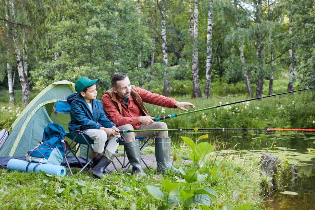 Full length portrait of loving father and son fishing together during camping trip by lake, copy space